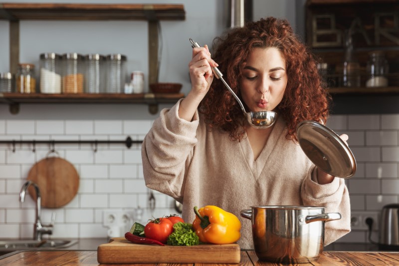 Gesunde Kochboxen - shutterstock 1399381838 Custom eine Frau kocht gesund und probiert die Suppe, die sie gemacht hat